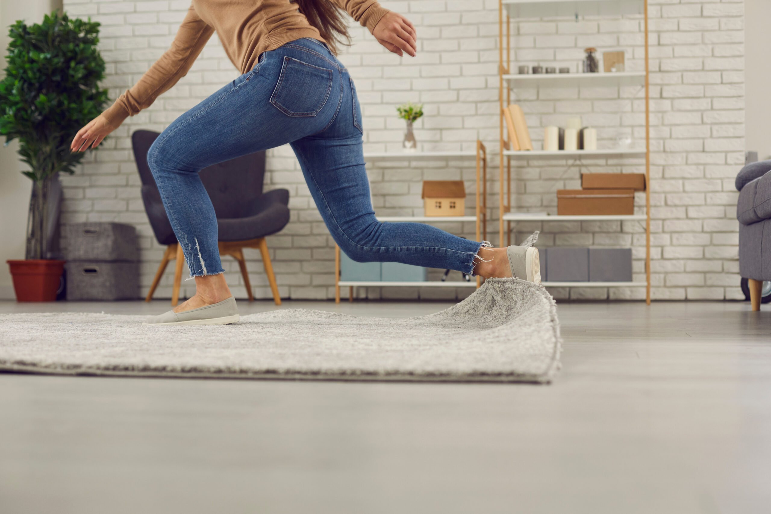 Close-up of a person in blue jeans tripping over the raised corner of a grey area rug in a brightly lit room.